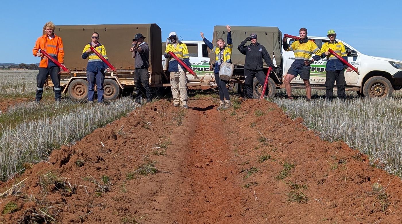 The planting crew standing along a ripped red-soil planting line with their utes and planting tools in a Wheatbelt paddock